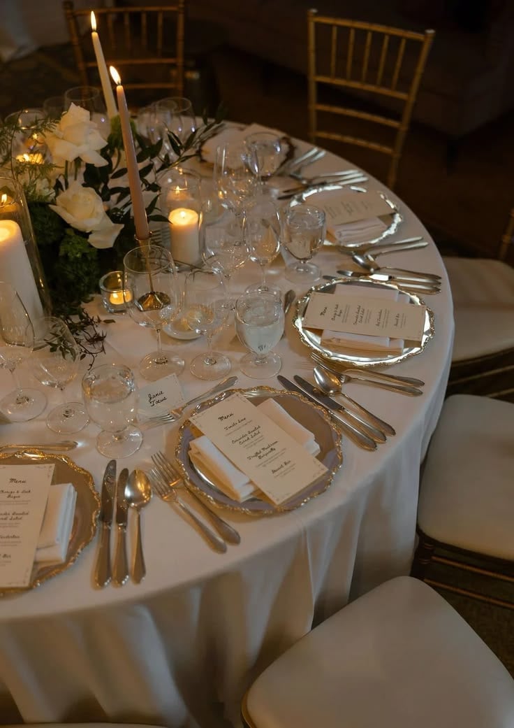 Elegant round dining table set for formal dinner with white linens, gold flatware, place cards, and floral centerpiece with candlelight