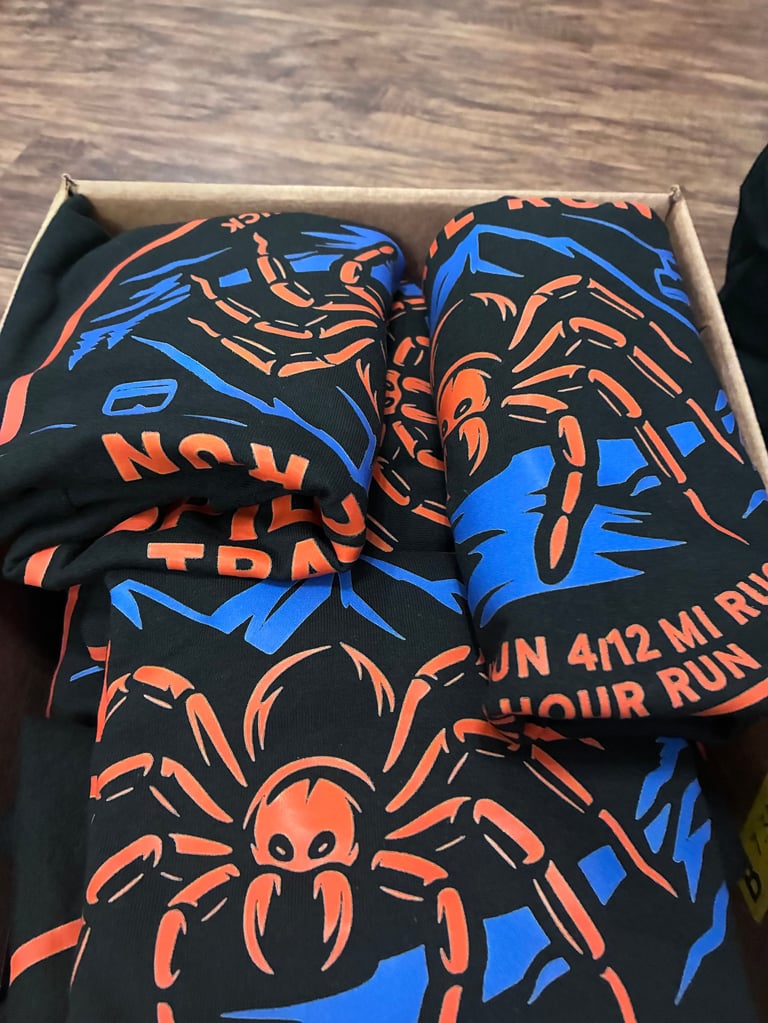Folded black t-shirts with bold orange and blue spider graphic designs and text in a cardboard box
