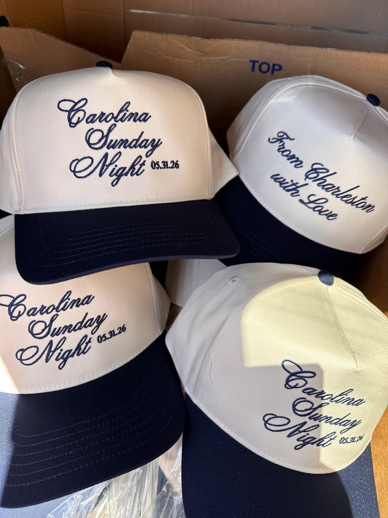 Collection of embroidered baseball caps with Carolina Sunday Night text and dates, white crown with navy bill in cardboard box