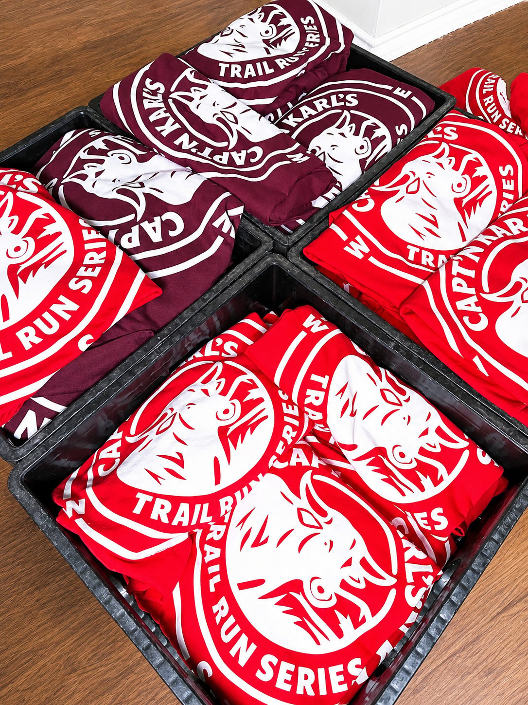 Stacked apparel featuring red and maroon Trail Series branded t-shirts with horse logos displayed in a black crate on wooden floor