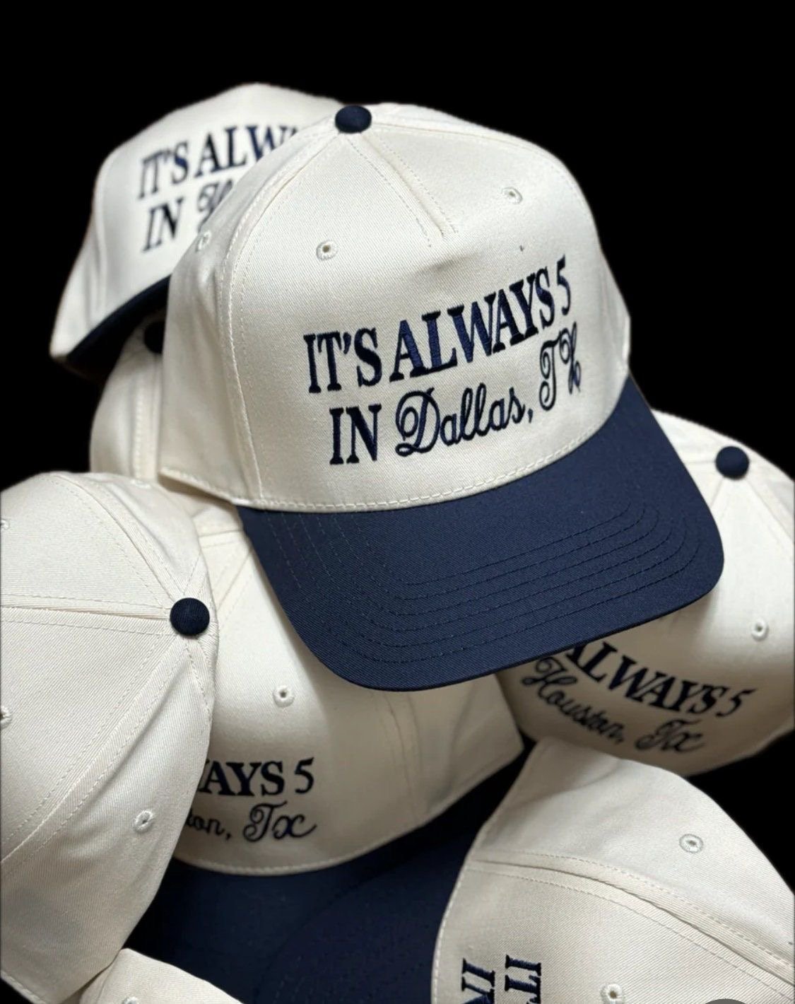 Stack of white and navy blue baseball caps with Its Always in Dallas, TX text printed on them