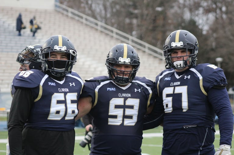Three football players in navy uniforms with gold accents standing together on the field, wearing helmets with face masks.