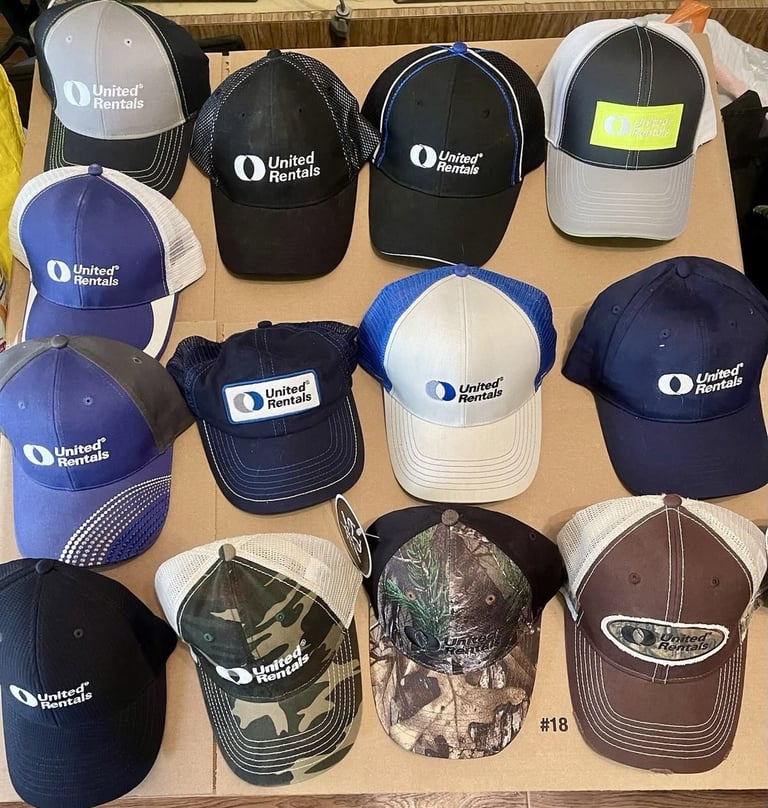 Collection of United Rentals branded baseball caps in various colors displayed on a tan surface, including black, white, blue, and camouflage styles.