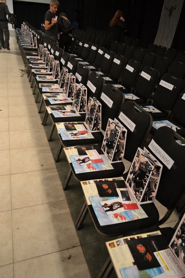 Long row of black chairs lined up with magazines on seats, person standing in background at what appears to be an indoor venue or event space