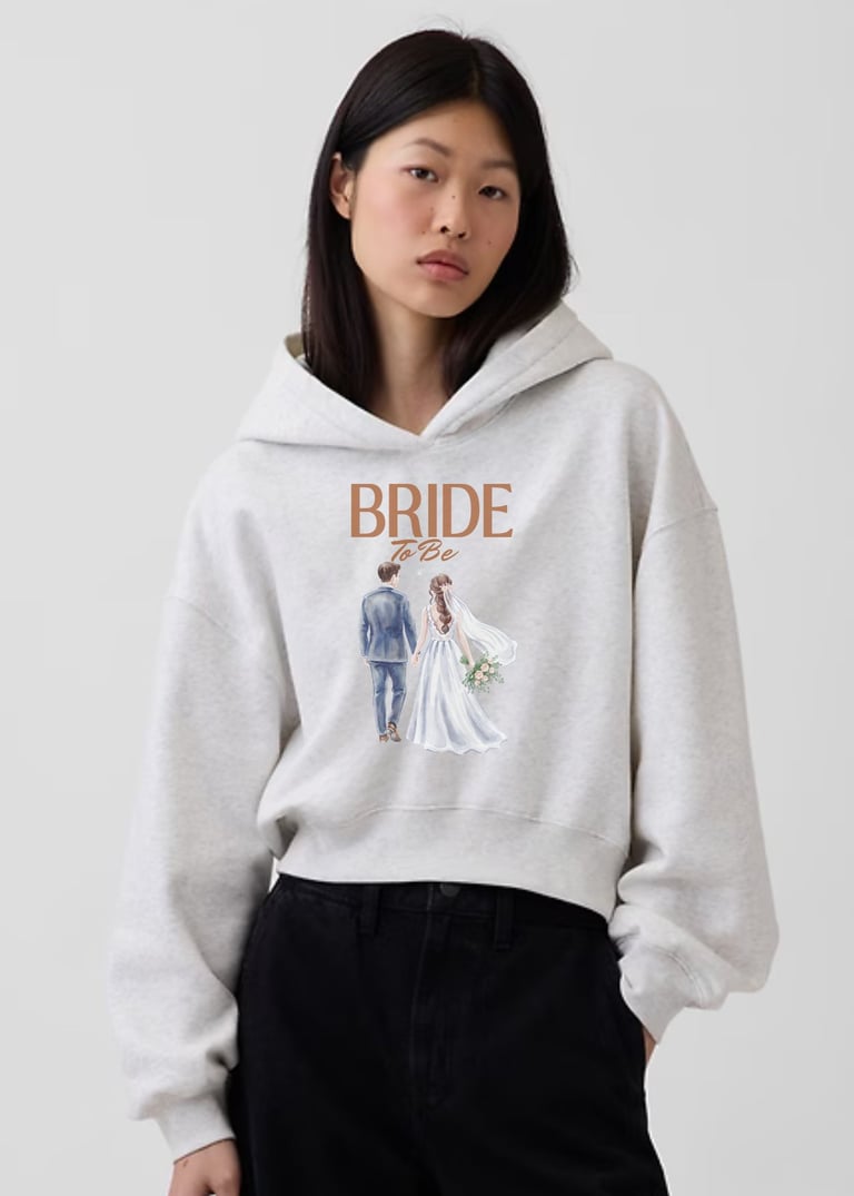 Woman wearing a light gray cropped hoodie with bride and groom illustration and black pants against a white background