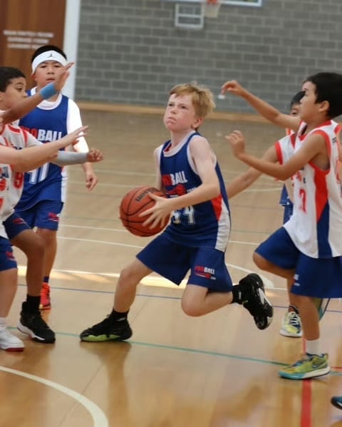 Young basketball player in blue jersey dribbling ball while being defended by opposing red team players during youth game in gymnasium