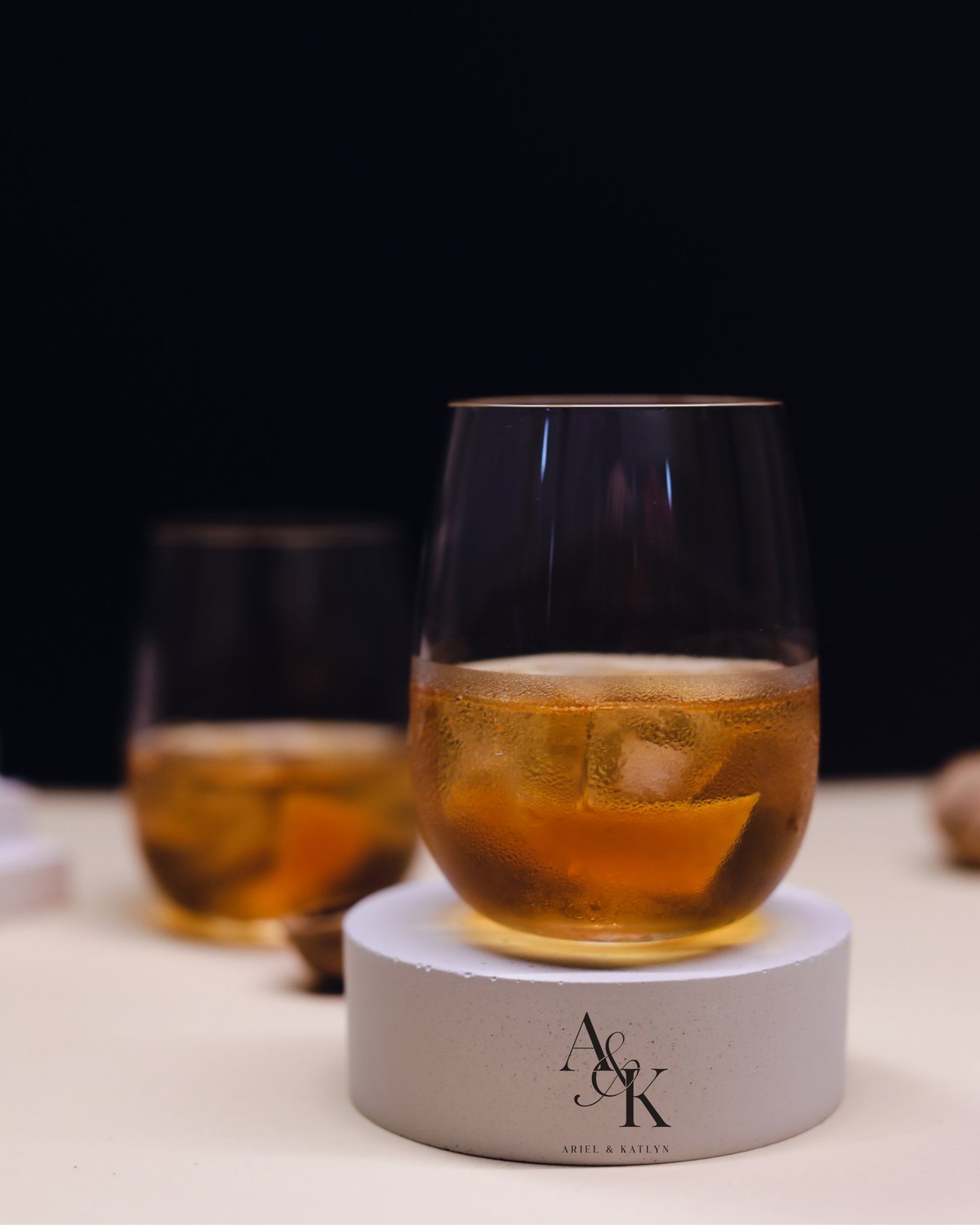 Two glasses of amber-colored whiskey with ice on white coasters with AK initials against a dark background