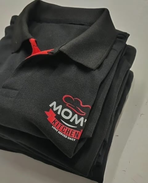 Black polo shirt neatly folded with red MOM KITCHEN logo and red tag on collar