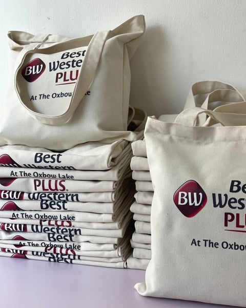 Collection of cream-colored canvas tote bags printed with Best Western Plus and Oxbow Lake branding, stacked and arranged on a surface